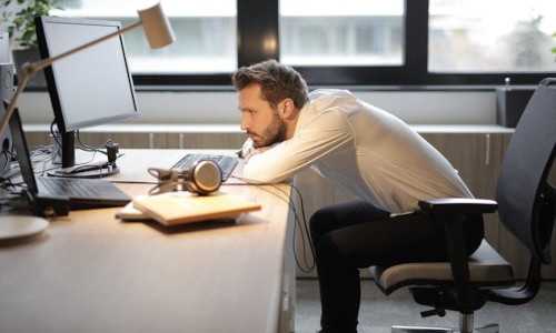 Zeigt einen Mann im Büro, der gelangweilt an einem Schreibtisch lümmelt und in seinen Laptop schaut.