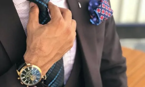 Zeigt einen Ausschnitt eines Mannes im braunen Anzug mit teurer Uhr, Krawatte und Einstecktuch